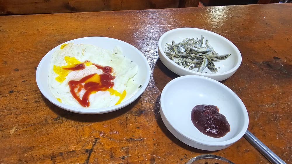 동래 오딧세이 포차 기본 안주인 계란후라이와 마른 멸치. 혼술하기 좋은 아늑한 나무 테이블 좌석 이미지.