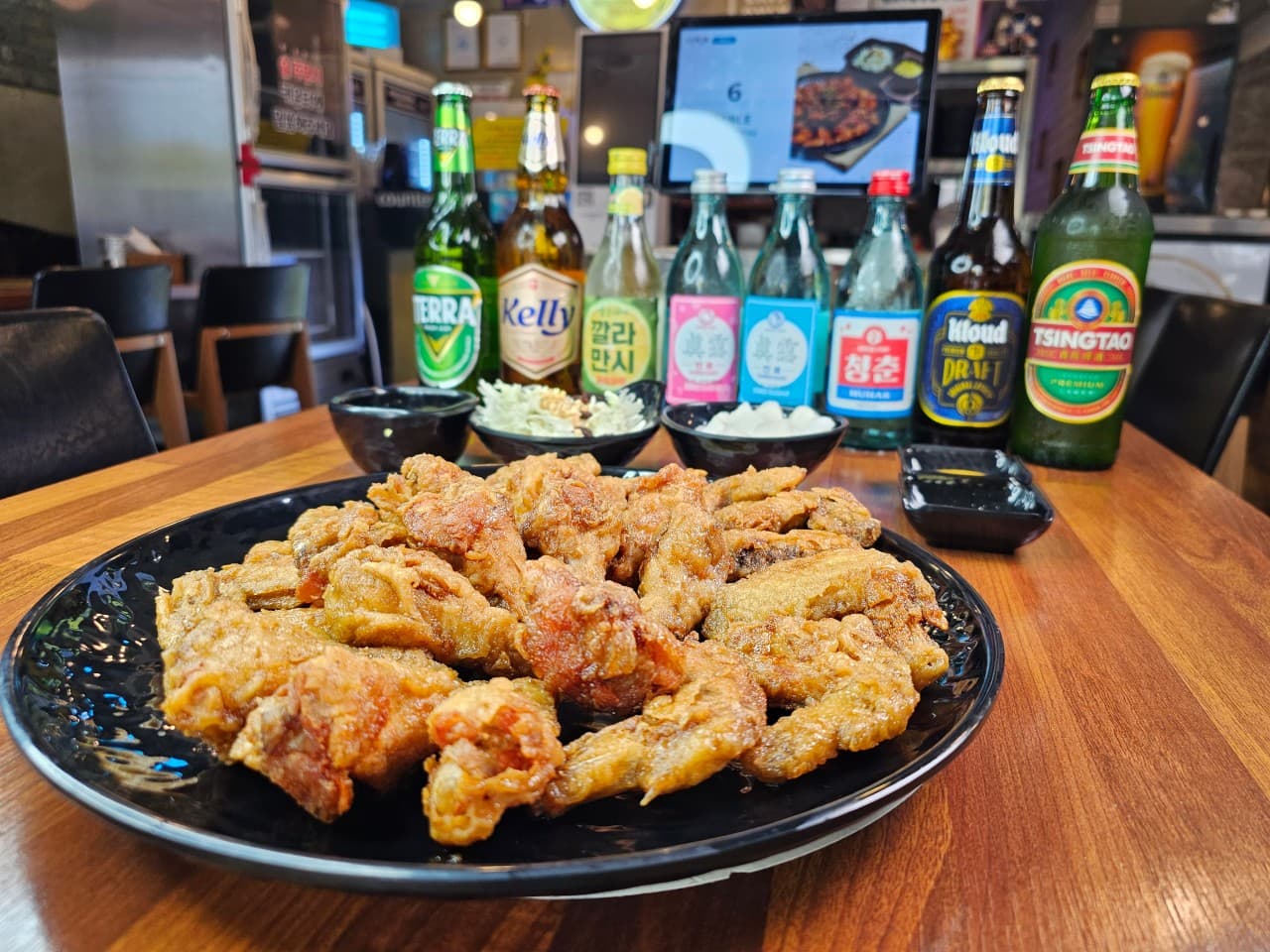 나무 테이블 위 검은 접시에 담긴 프라이드 치킨과 그 뒤로 일렬로 늘어선 다양한 종류의 병맥주와 소주