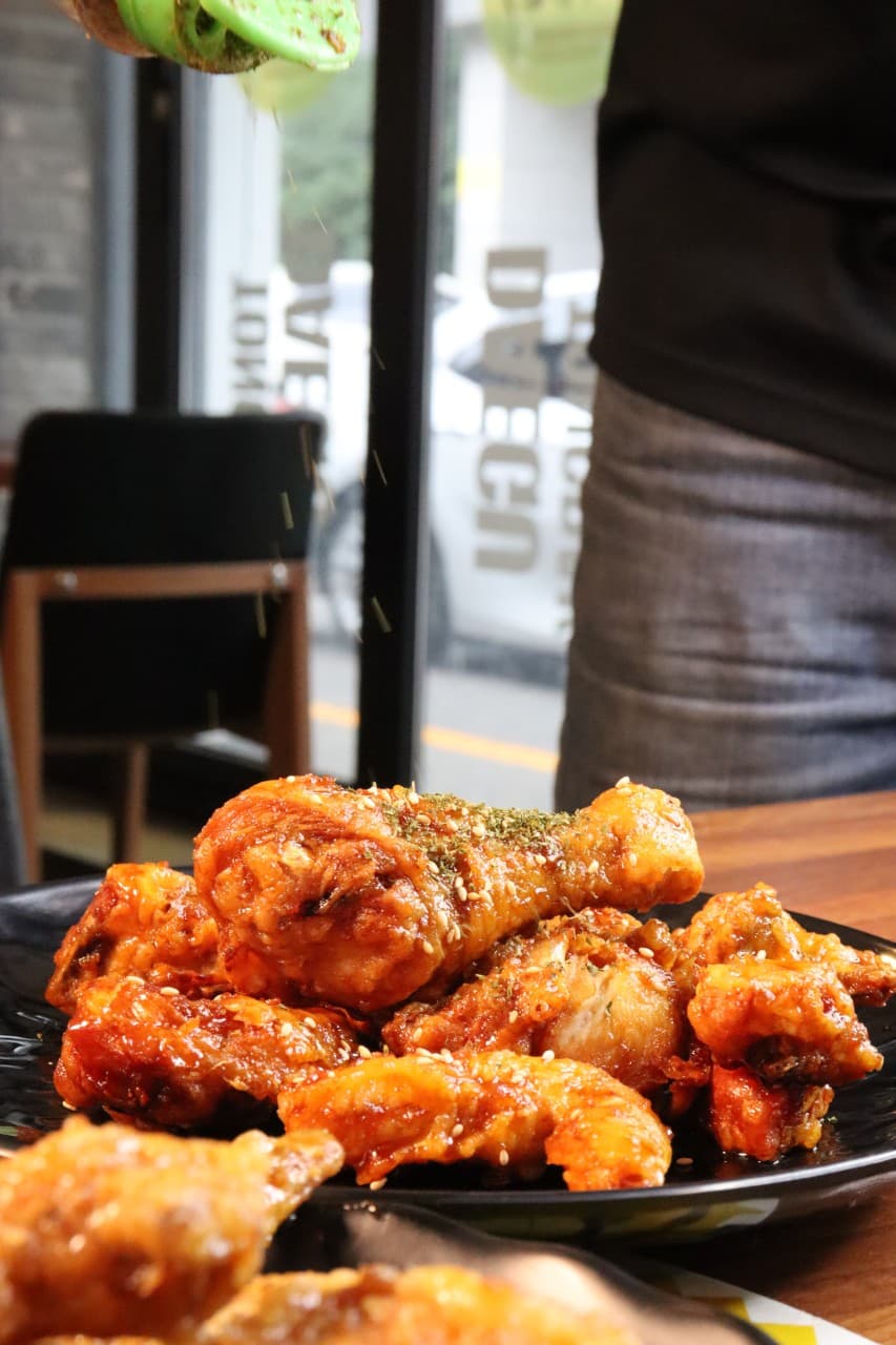 검은 접시에 담긴 붉은 양념 치킨 위로 뿌려지는 파슬리 가루. 창가 쪽 목재 테이블에 놓인 1인용 식사 구성