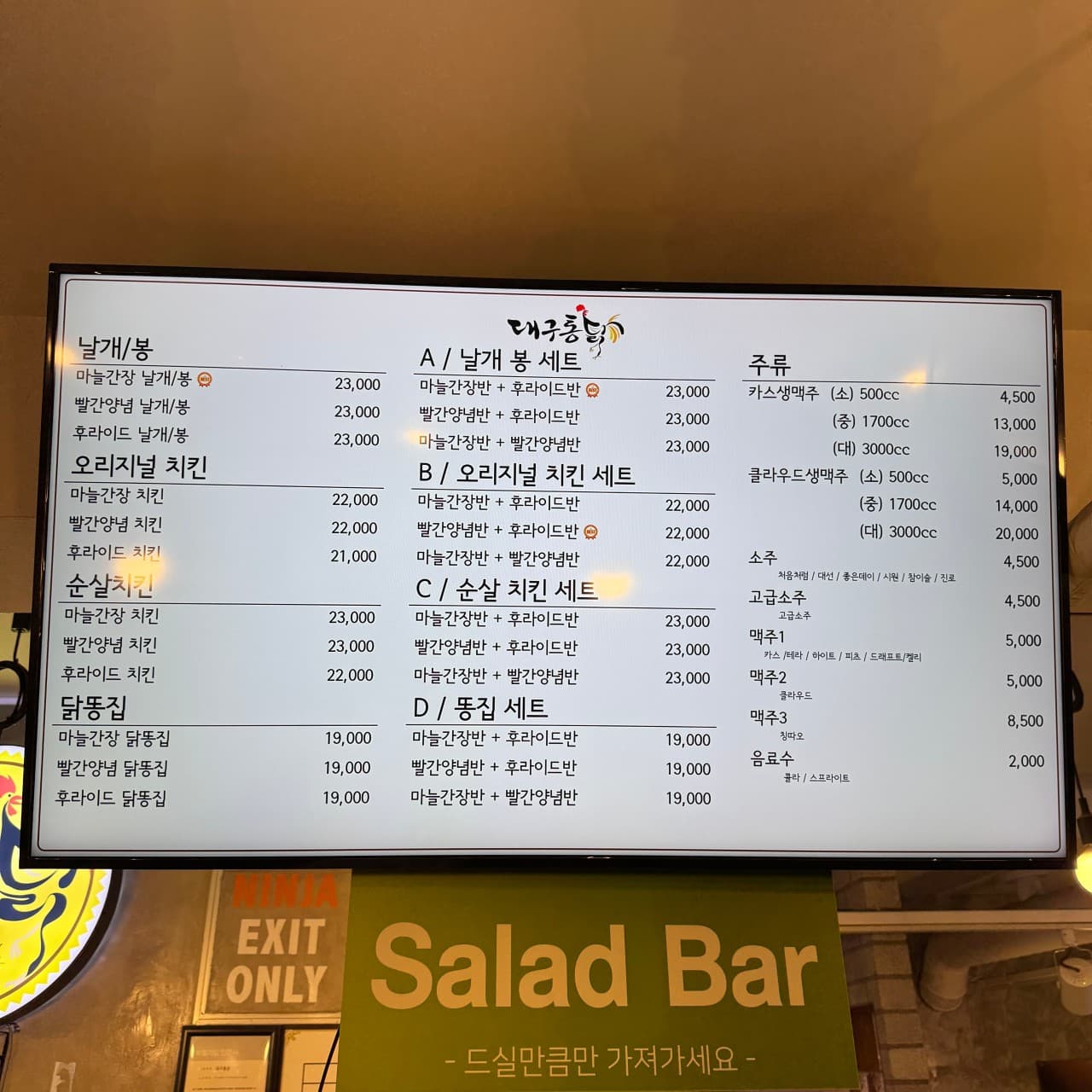치킨 세트와 생맥주 가격이 상세히 적힌 밝은 디지털 메뉴판 및 하단 셀프 샐러드바 안내 문구가 보이는 매장 내부