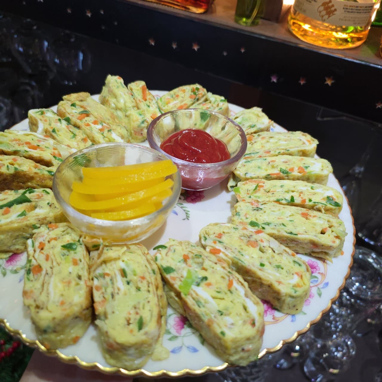 어두운 바 테이블 위 꽃무늬 접시에 원형으로 놓인 야채 계란말이와 케첩, 노란 단무지가 담긴 작은 유리 용기