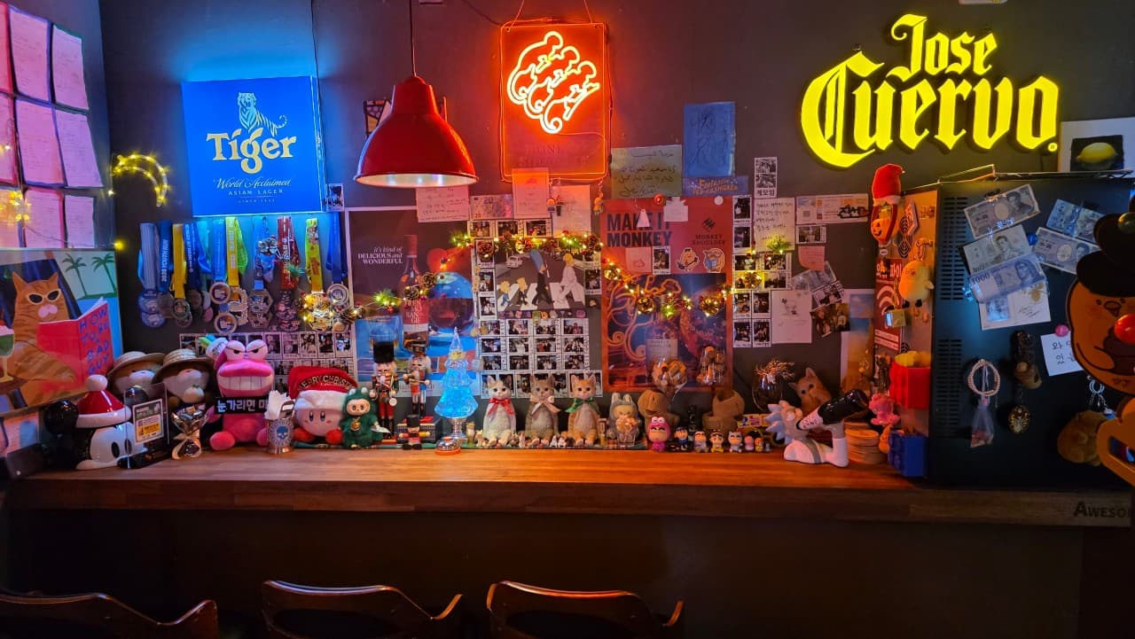푸른색과 노란색 네온 사인 아래 수많은 피규어와 포스터로 장식된 1인용 목재 카운터 좌석