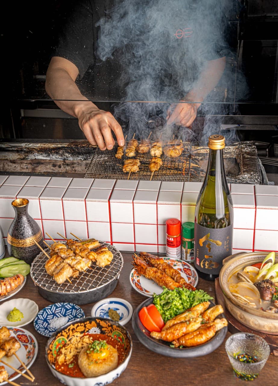 연기가 나는 화로에서 꼬치를 굽는 요리사와 화이트 타일 카운터 위 닭꼬치, 사케 병, 전골 등 1인용 식기가 놓인 상차림