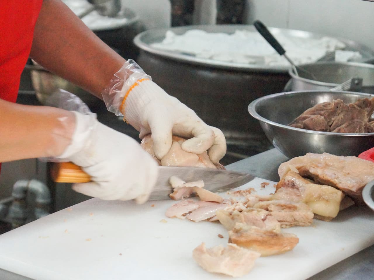 흰색 도마 위에서 면장갑을 끼고 일정한 두께로 써는 수육과 주방 조리대 위 스테인리스 용기