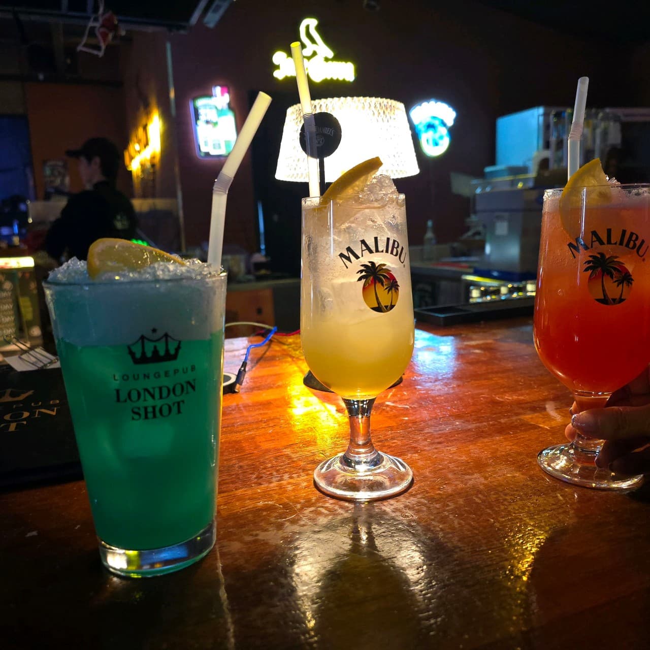 나무 질감의 카운터 위 레몬과 빨대가 꽂힌 세 종류의 칵테일 잔. 네온 사인과 조명이 비치는 어두운 실내의 바 테이블 전경