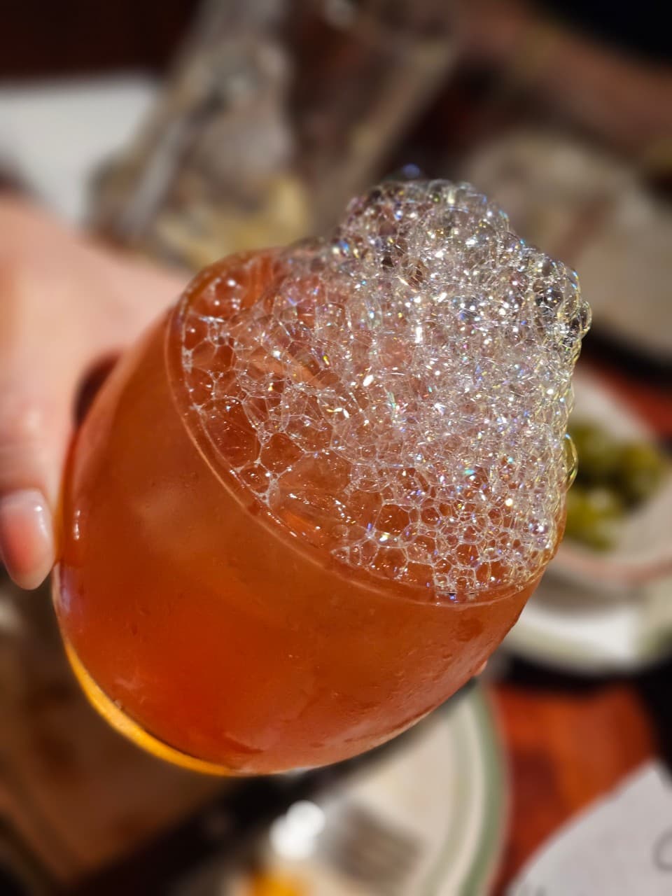 조명 아래 무지개빛 광택이 도는 거품이 쌓인 하이볼 잔을 든 손과 바 테이블 위 음식 접시