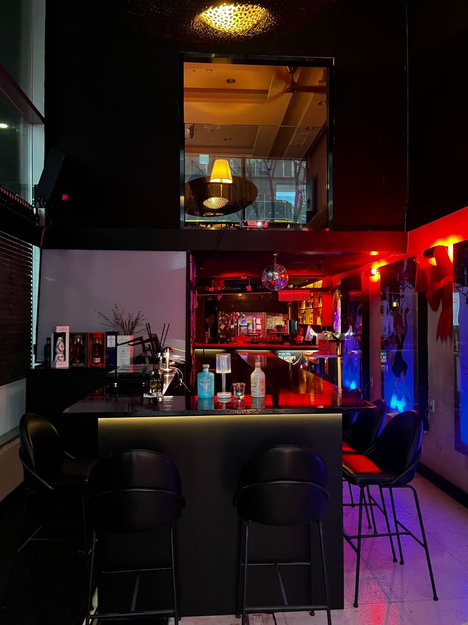붉은 조명이 비치는 어두운 실내에 배치된 검은색 바 카운터와 높은 의자들. 테이블 위 술병과 작은 램프가 놓인 전경