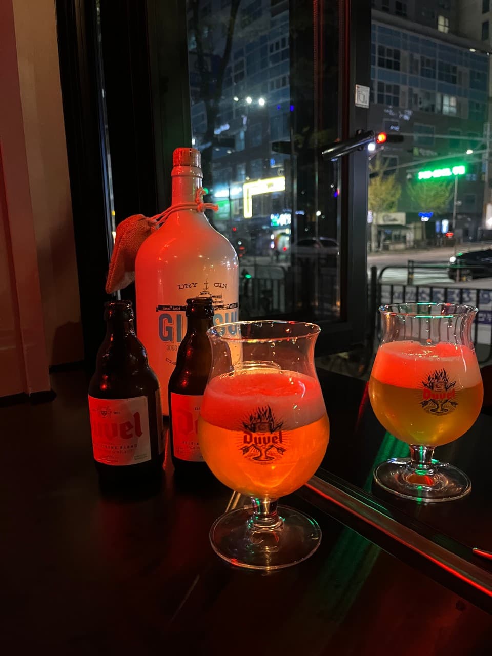 어두운 밤거리 야경이 보이는 창가 바 테이블 위 붉은 조명을 받는 맥주 전용 잔과 맥주병 및 진 보틀