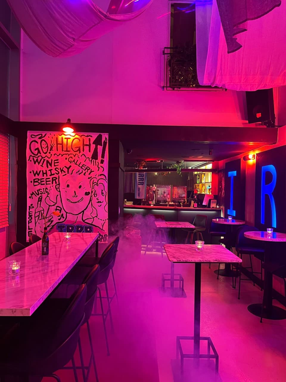 보랏빛 조명 아래 대리석 상판의 높은 테이블과 1인용 의자들. 바닥에 깔린 자욱한 연기와 벽면의 팝아트 포스터.