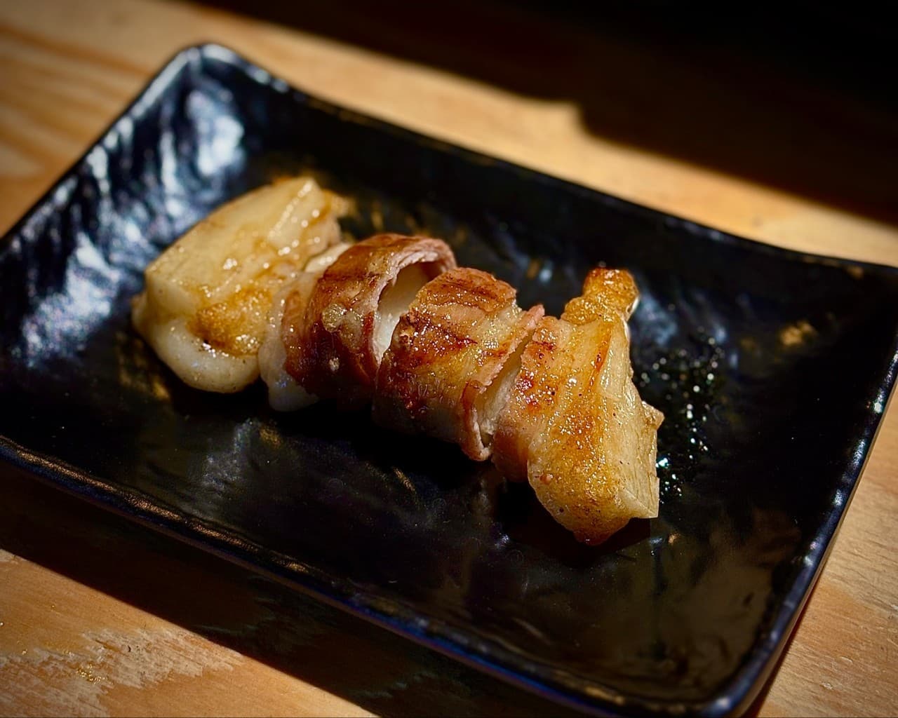 나무 소재의 카운터 테이블 위 검은색 사각 접시에 담긴 노릇하게 구워진 1인용 고기 꼬치구이