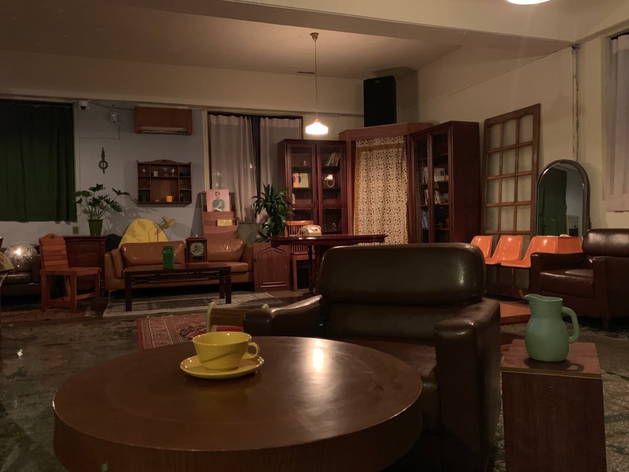 목재 원형 테이블 위 노란 찻잔과 1인용 가죽 소파. 전구 조명 아래 나무 책장과 가죽 소파, 주황색 플라스틱 의자가 독립적으로 배치된 공간