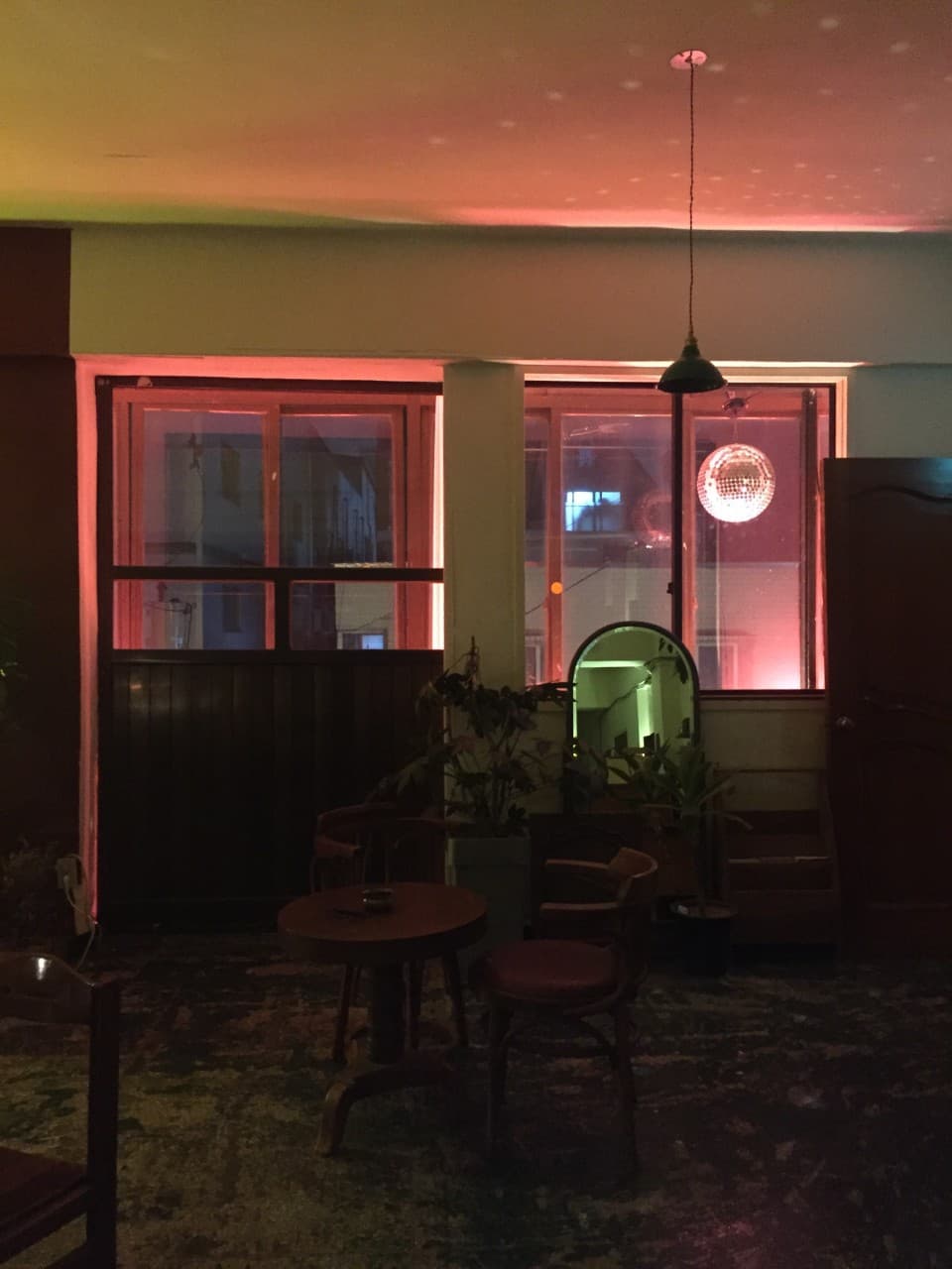 붉은 조명이 비치는 어두운 실내에 놓인 작은 원형 목재 테이블과 의자, 창가의 미러볼과 식물들