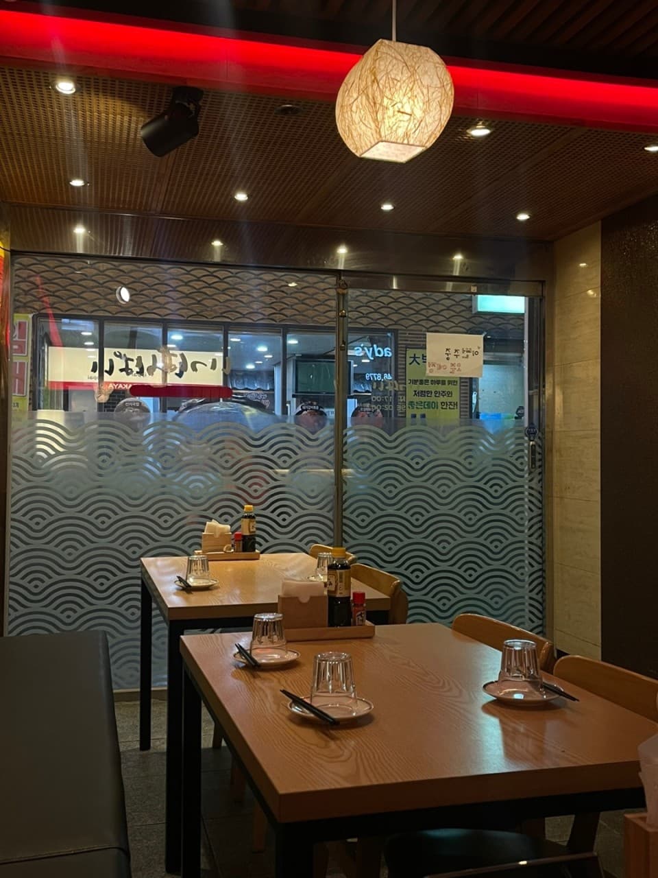 붉은 네온 조명 아래 컵과 접시가 세팅된 나무 테이블. 물결무늬 시트지가 부착된 유리문 앞 2인용 좌석이 배치된 매장 내부 전경