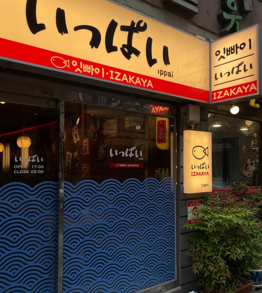 노란색 조명 간판과 푸른 파도 무늬가 부착된 유리창. 내부의 종이 등불과 포스터가 보이는 일본식 선술집 건물 입구 전경