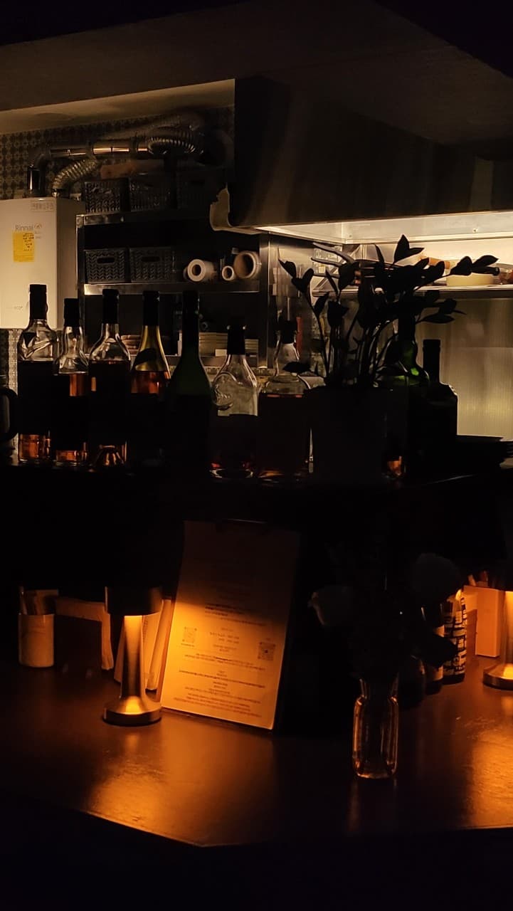 어두운 조명 아래 스탠드와 메뉴판이 놓인 바 테이블 전경과 뒤편에 나열된 주류 병들