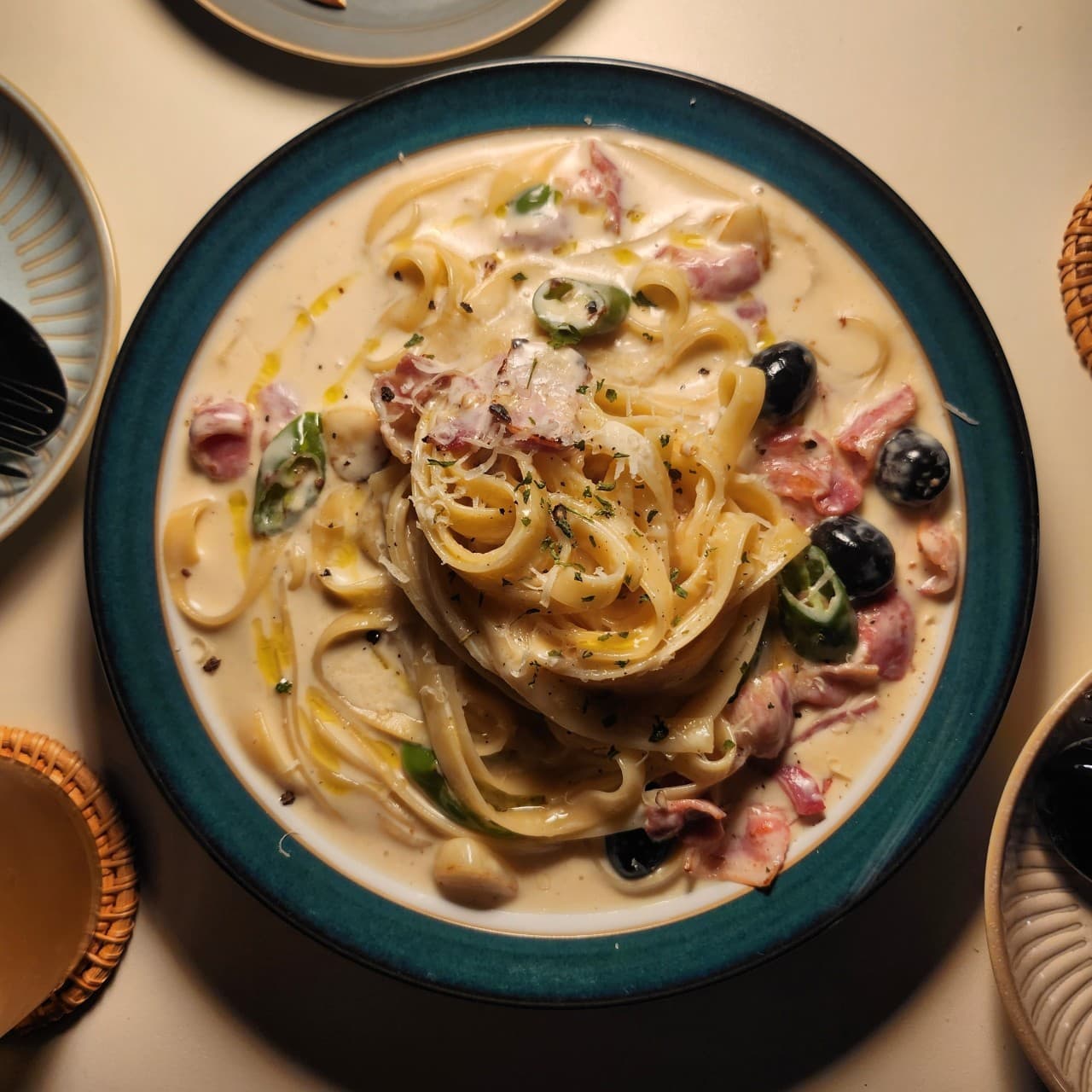 청록색 테두리 접시에 담긴 베이컨과 올리브를 곁들인 크림 파스타. 조명 아래 1인용 식기와 음료가 놓인 테이블 전경