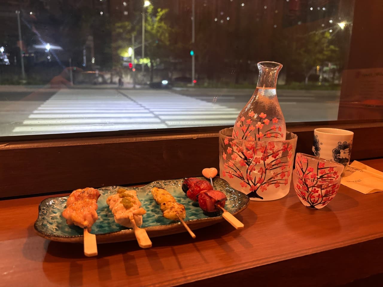 밤 거리 횡단보도가 보이는 창가 목재 카운터 위 꼬치 네 점이 담긴 접시와 벚꽃 무늬 술병 세트