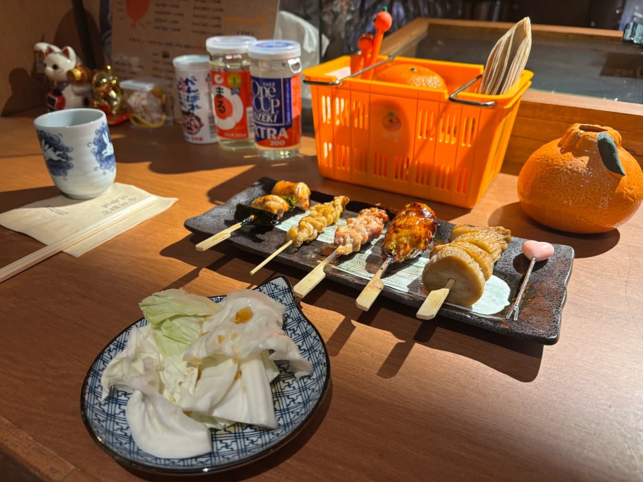 밝은 조명이 비치는 목재 다찌석 위 꼬치구이 5종과 양배추 접시, 컵 사케와 주황색 소품이 놓인 1인용 차림