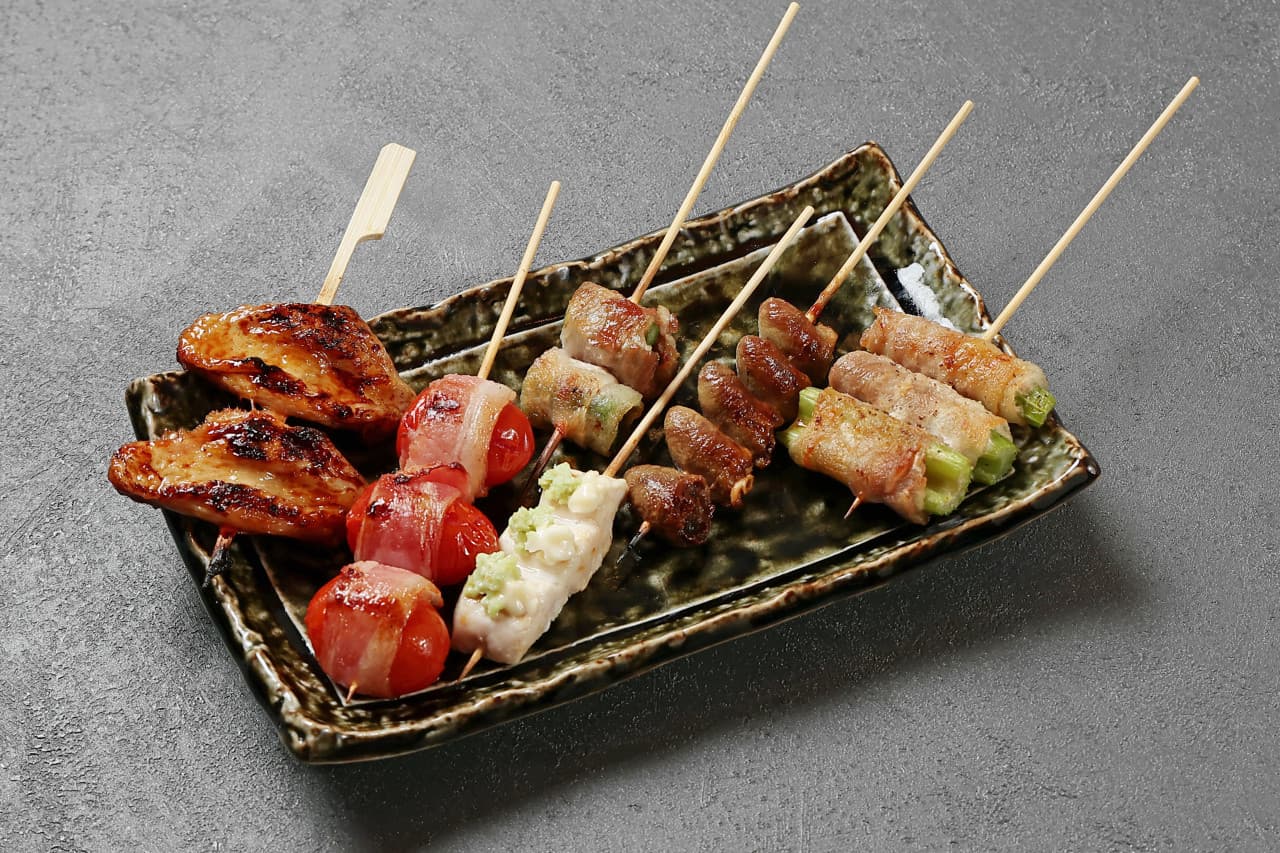 거친 질감의 회색 테이블 위 도자기 접시에 담긴 닭날개, 토마토 베이컨 말이, 염통 등 1인분 분량의 다양한 꼬치구이 모듬
