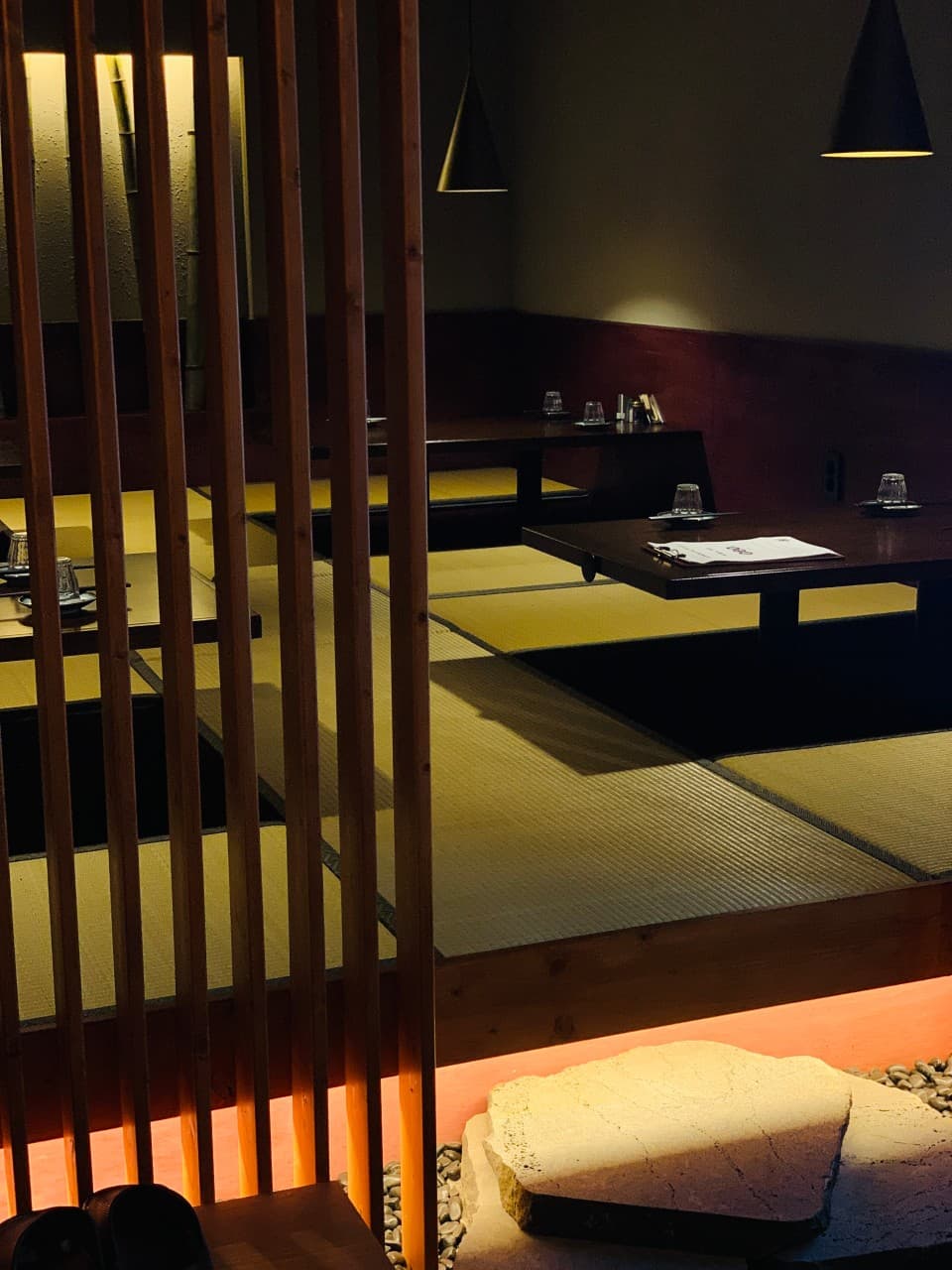 목재 살 너머로 보이는 다다미 좌식 테이블과 1인용 식기 세팅, 하단부의 오렌지색 조명과 자갈
