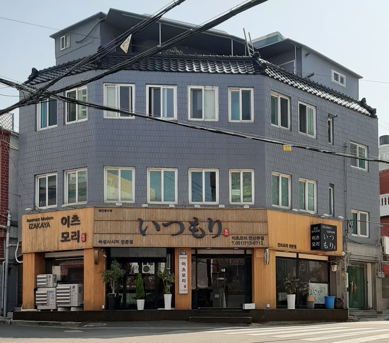 목재 외벽과 통유리창으로 구성된 1층 일식 주점. 회색 타일 건물 코너에 위치하며 입구 주변에 작은 화분들이 놓인 외부 전경