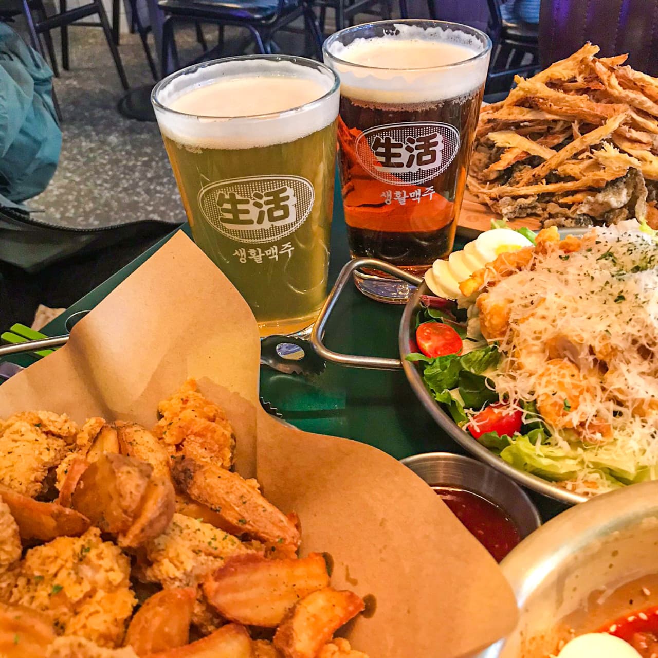 거품이 올라온 맥주 두 잔과 종이 위 후라이드 치킨, 감자튀김, 치즈 가루를 듬뿍 얹은 샐러드와 마른 안주가 차려진 테이블 위 전경