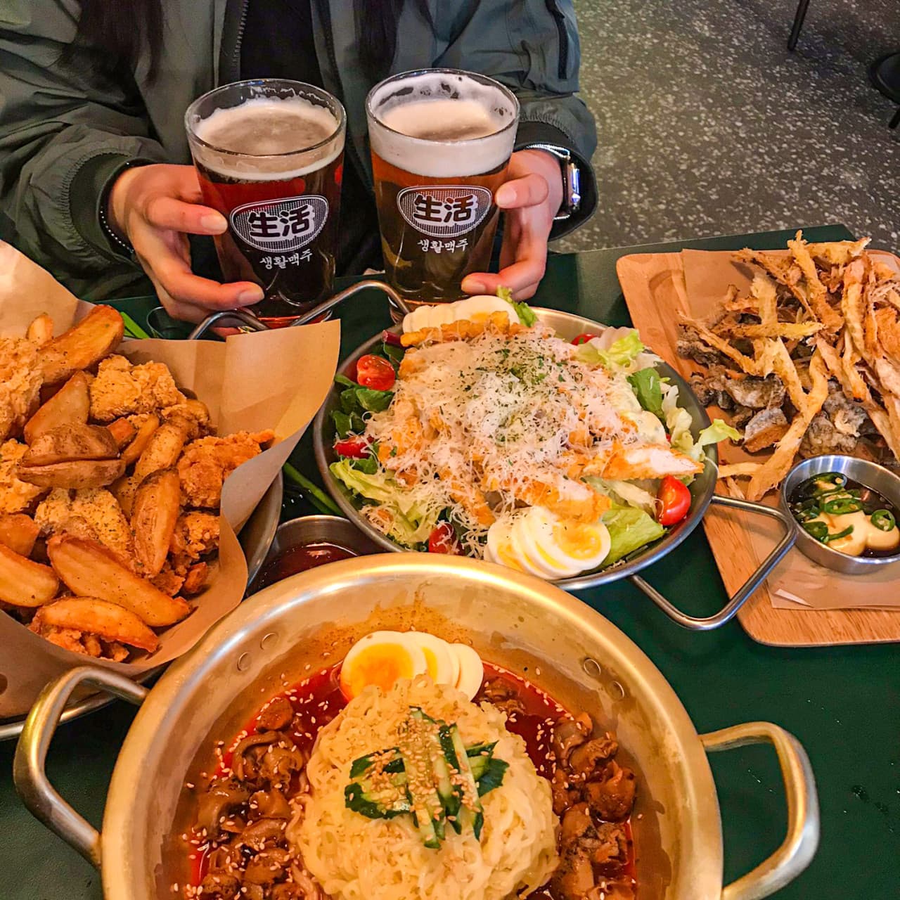 어두운 조명 아래 양은 냄비에 담긴 비빔면과 조각 치킨, 샐러드, 나무 트레이 위 마른안주와 거품이 올라온 맥주잔