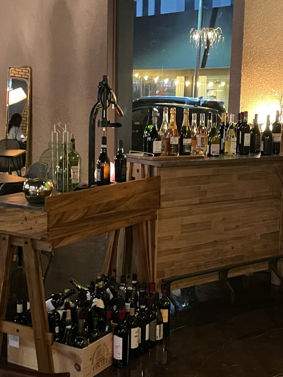 어두운 실내 조명 아래 나무 카운터 위 와인병들과 바닥의 빈 병들, 창문 너머로 보이는 밤의 거리와 주차된 차량