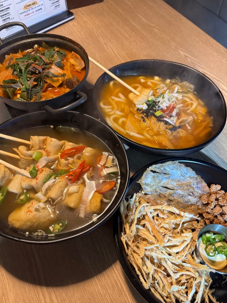 나무 질감 테이블 위 꼬치어묵탕, 김치우동, 붉은 전골 냄비와 소스를 곁들인 마른안주가 놓인 상차림