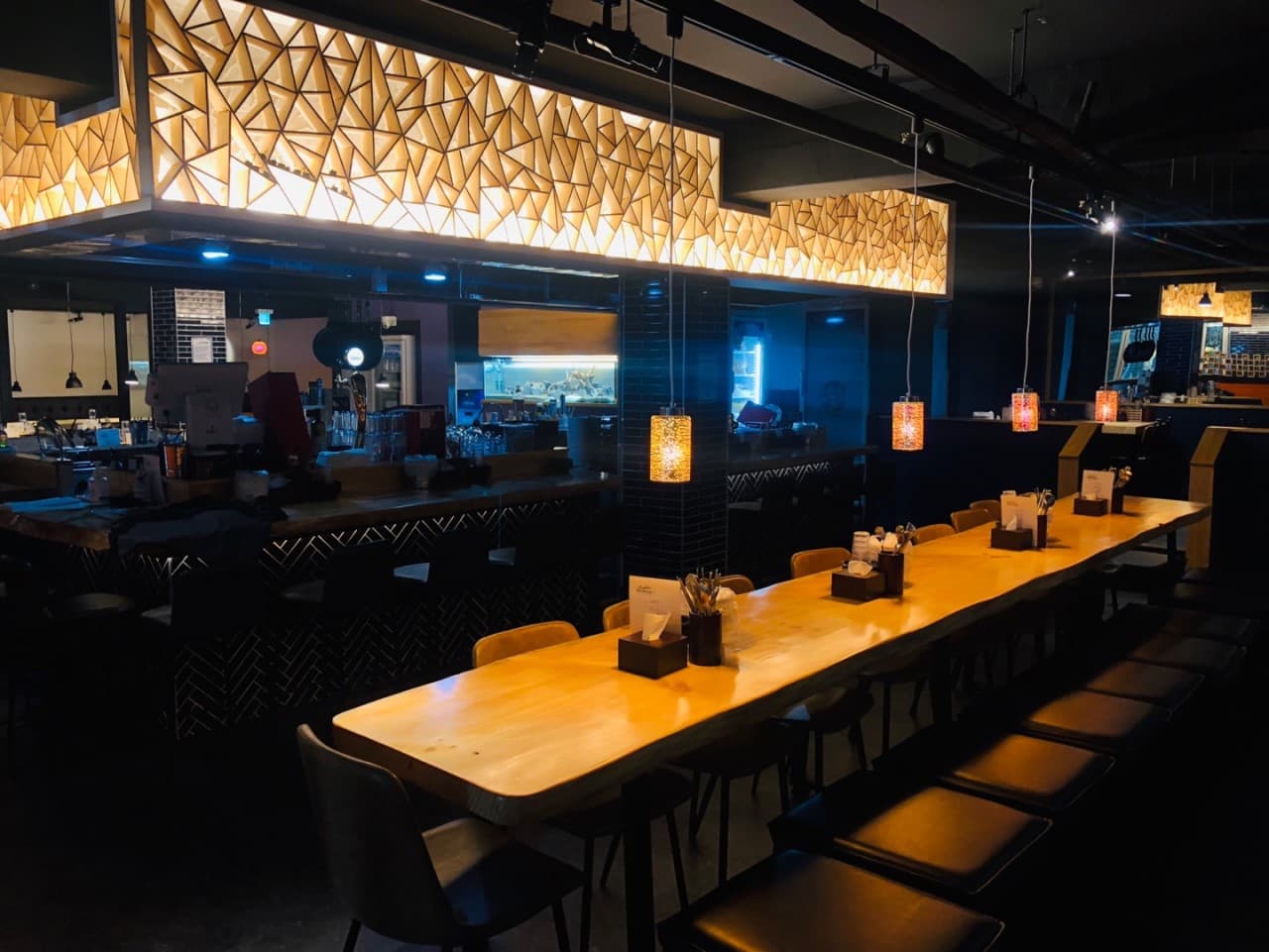 어두운 배경의 기하학적 천장 조명 아래 길게 배치된 원목 바 테이블과 나란히 정렬된 1인용 의자들