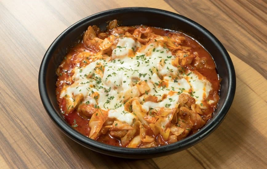 목재 테이블 위 1인용 검은 그릇에 담긴 붉은 양념의 볶음 요리와 녹아내린 하얀 치즈 및 파슬리