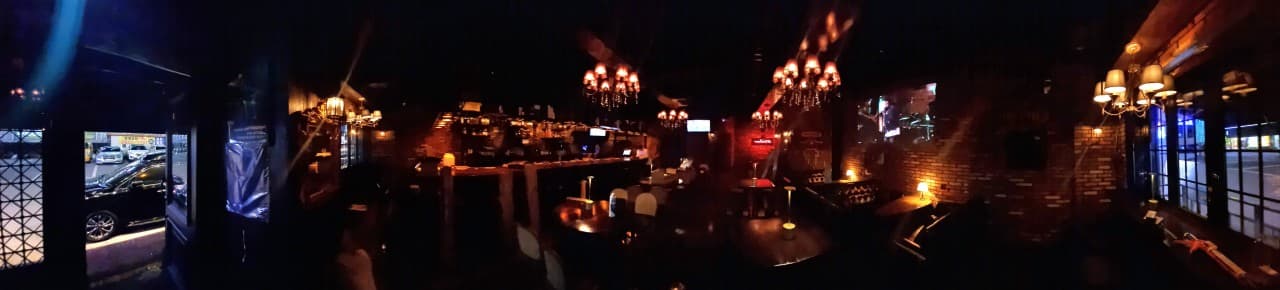 어두운 조명 아래 긴 목재 바 카운터와 1인용 의자들이 배치된 내부. 벽돌 벽면을 따라 낮은 조도의 샹들리에와 조명들이 켜진 전경.