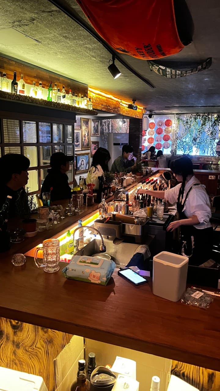 노란 간접 조명이 비치는 긴 목재 카운터 테이블과 띄엄띄엄 배치된 좌석에 앉은 손님 및 음료 제조 직원