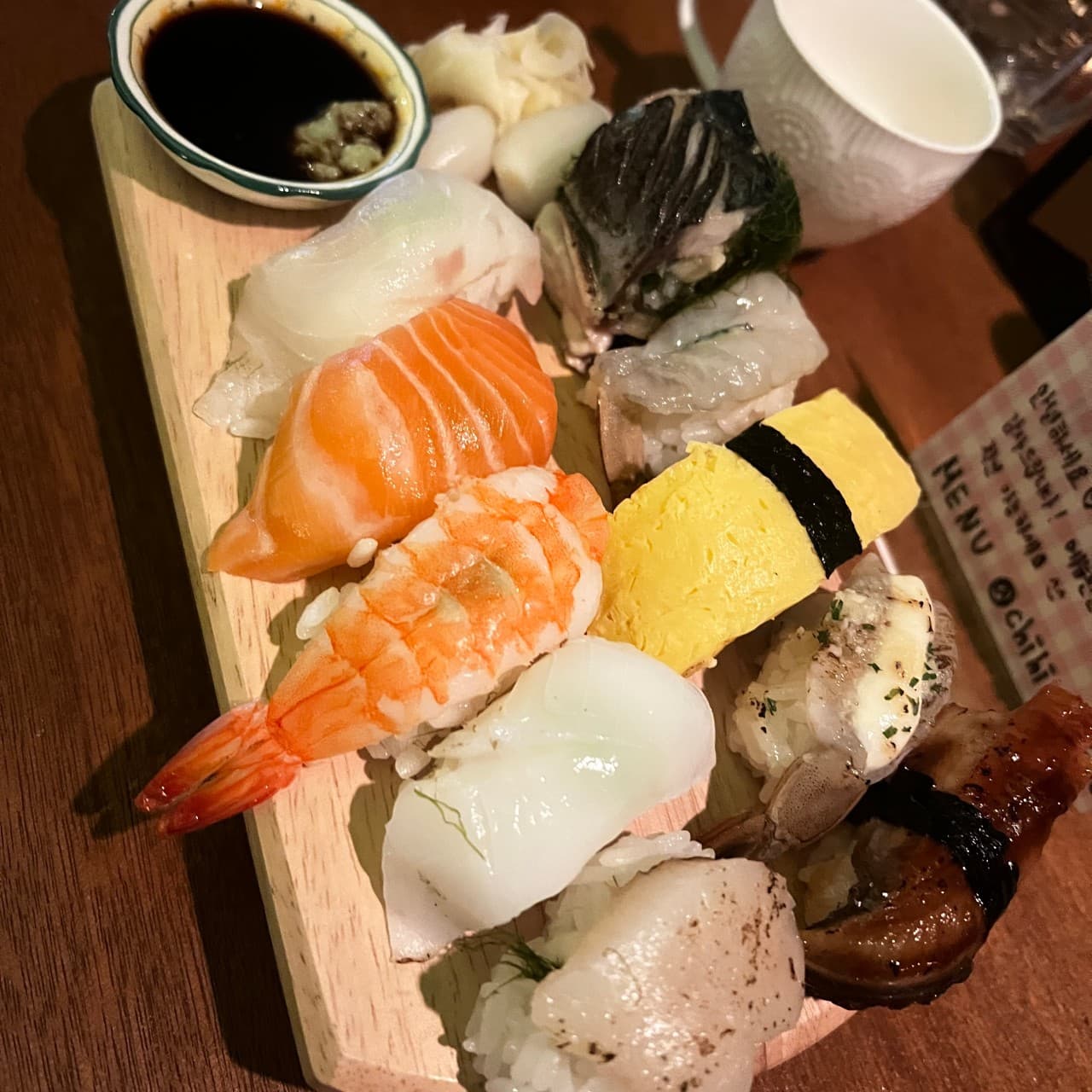 어두운 나무 테이블 위 1인용 나무 도마에 놓인 연어와 새우 등 모둠 초밥, 간장 종지와 하얀 잔