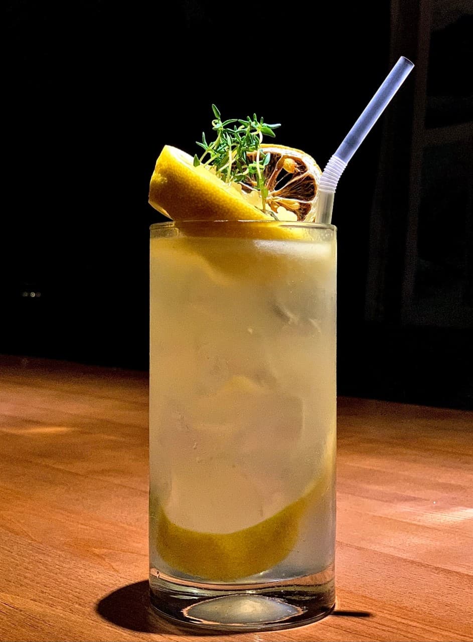 어두운 배경 속 나무 다찌석 위 레몬 조각과 허브가 장식된 얼음 가득한 하이볼 잔과 빨대