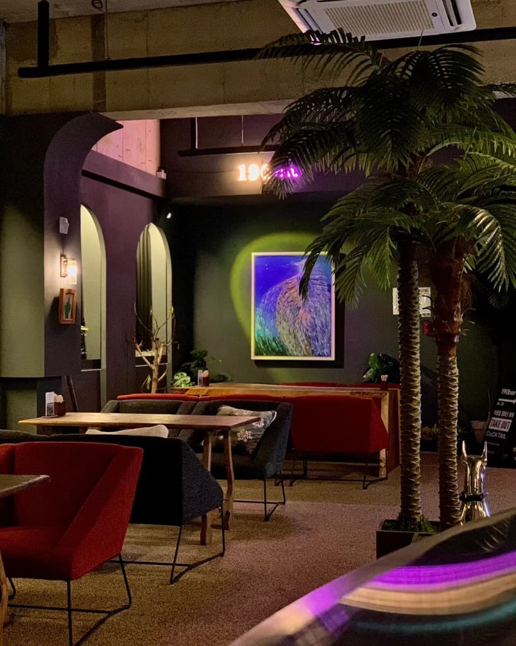 어두운 조명 아래 1인용 소파와 목재 테이블이 배치되어 있고 대형 야자수와 벽면 액자가 조명을 받는 전경