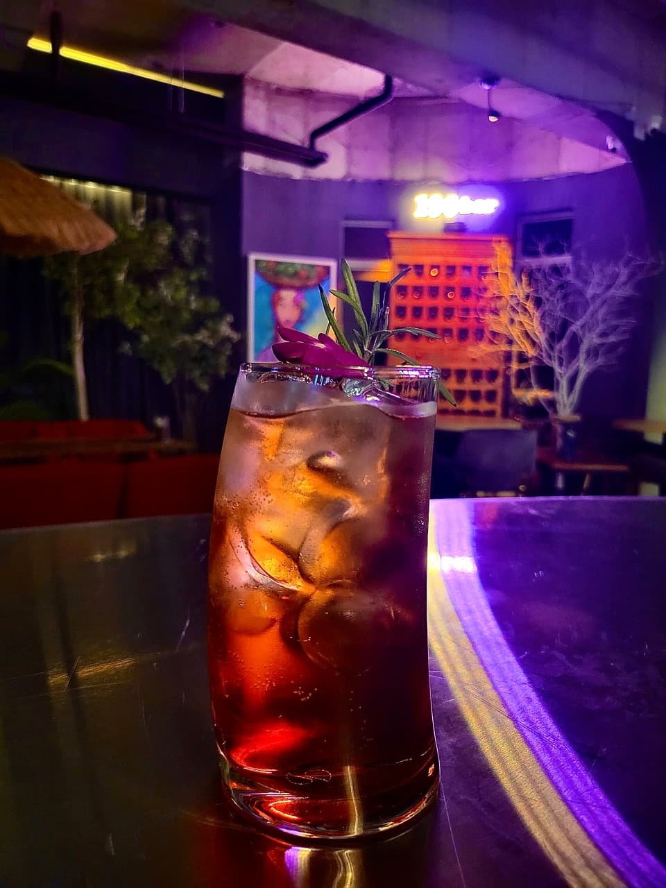 보라색 조명이 비치는 어두운 바 테이블 위 꽃과 로즈마리가 얹어진 칵테일 잔과 가득한 얼음