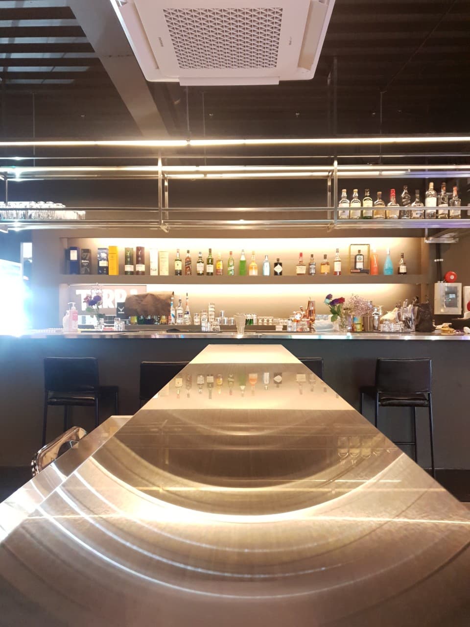 은은한 빛이 반사되는 긴 금속 카운터 테이블과 벽면 진열장에 늘어선 다양한 주류 병들, 그 앞에 놓인 1인용 검은색 의자