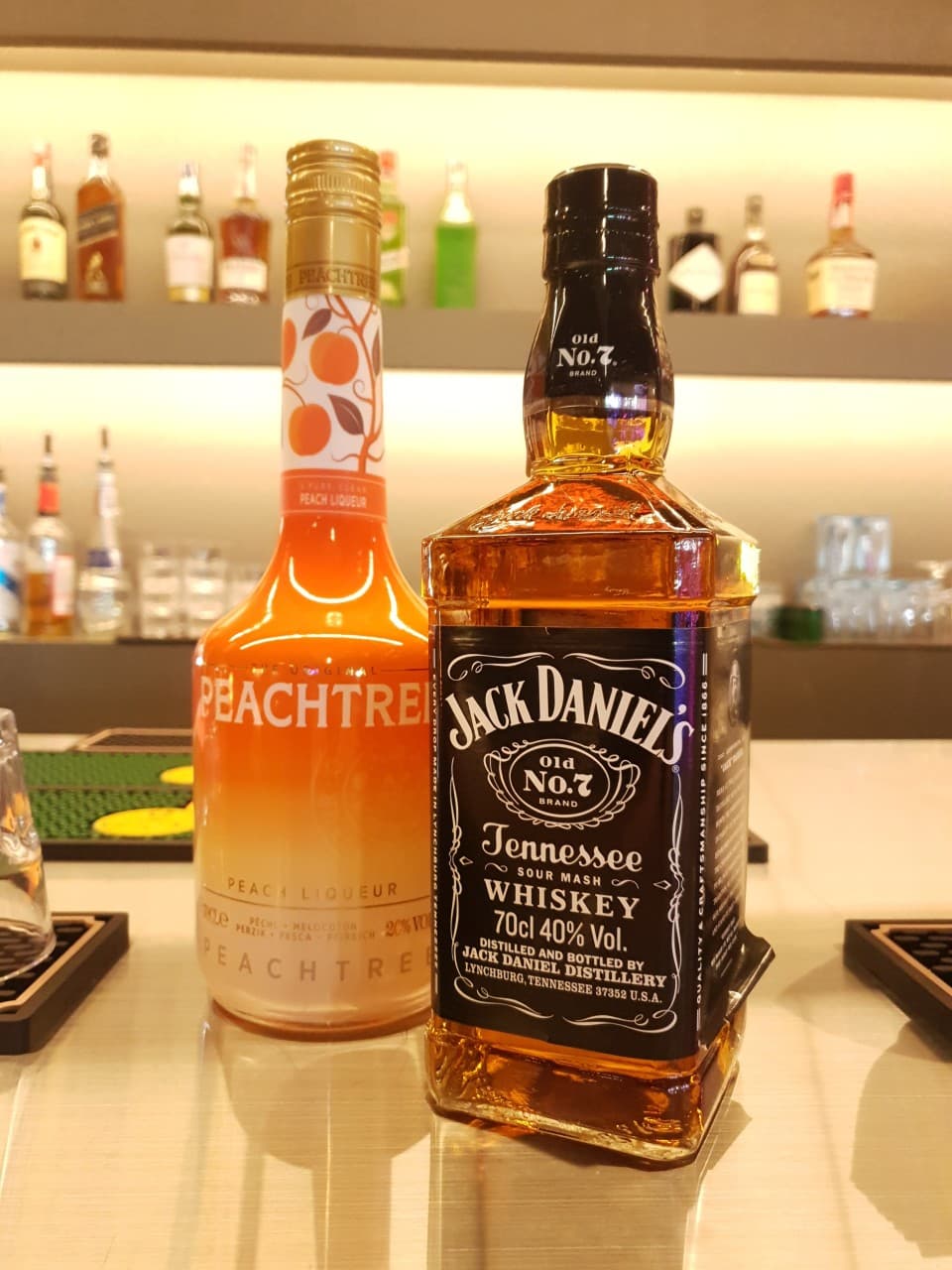 은은한 노란 조명이 비치는 바 테이블 위 잭다니엘 위스키와 피치트리 리큐르 병 그리고 배경에 진열된 다양한 술병들