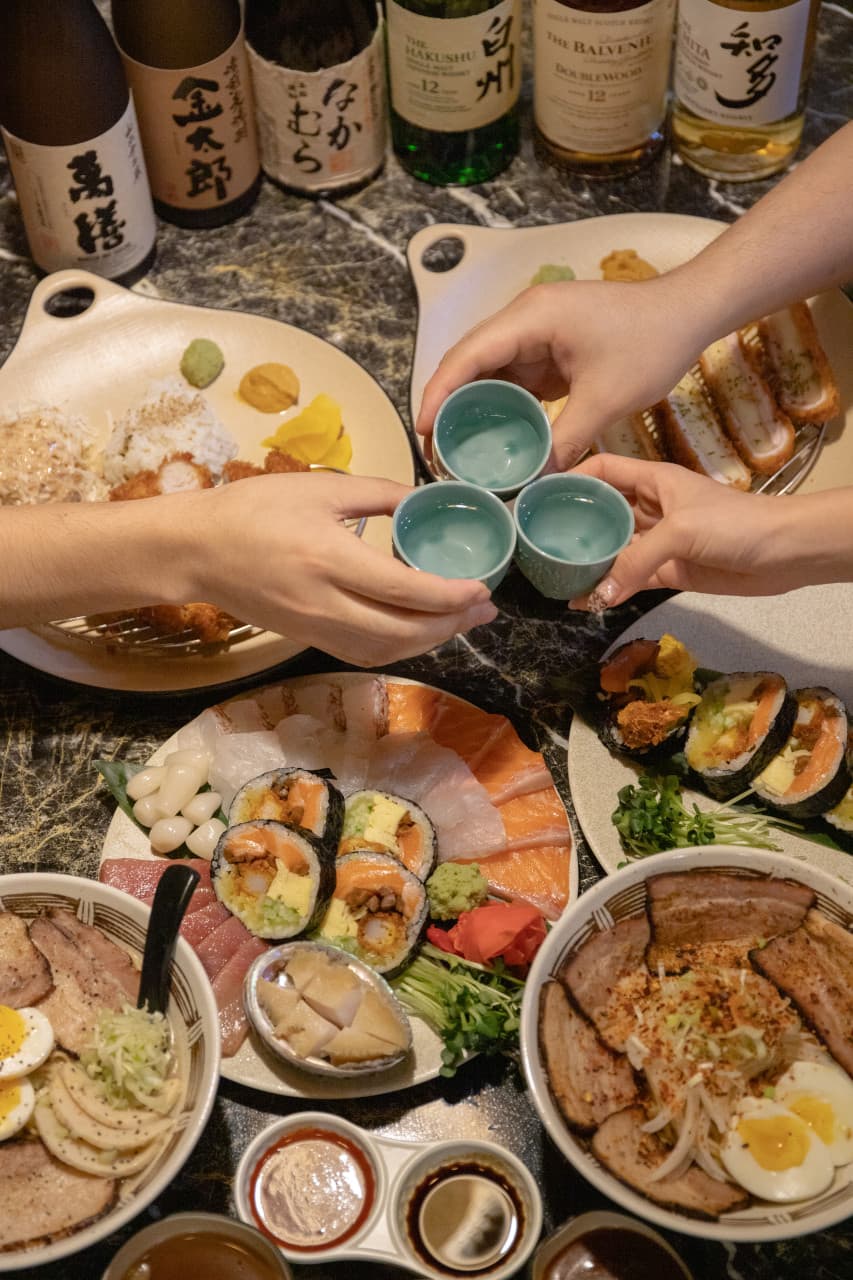 사시미와 후토마키, 라멘이 차려진 어두운 대리석 테이블 위 푸른 술잔을 맞대는 모습과 배경에 나열된 술병들