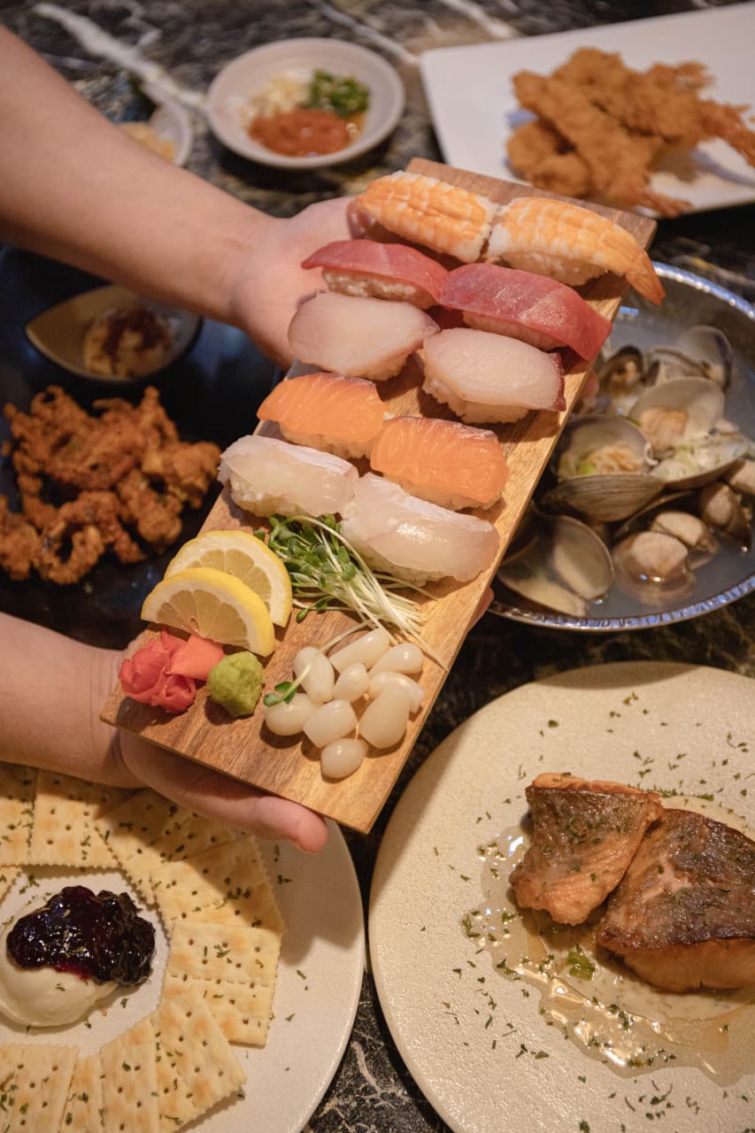 목재 트레이에 담긴 모둠 초밥과 연어 구이, 바지락 술찜, 크래커가 차려진 어두운 톤의 대리석 테이블 위 상차림