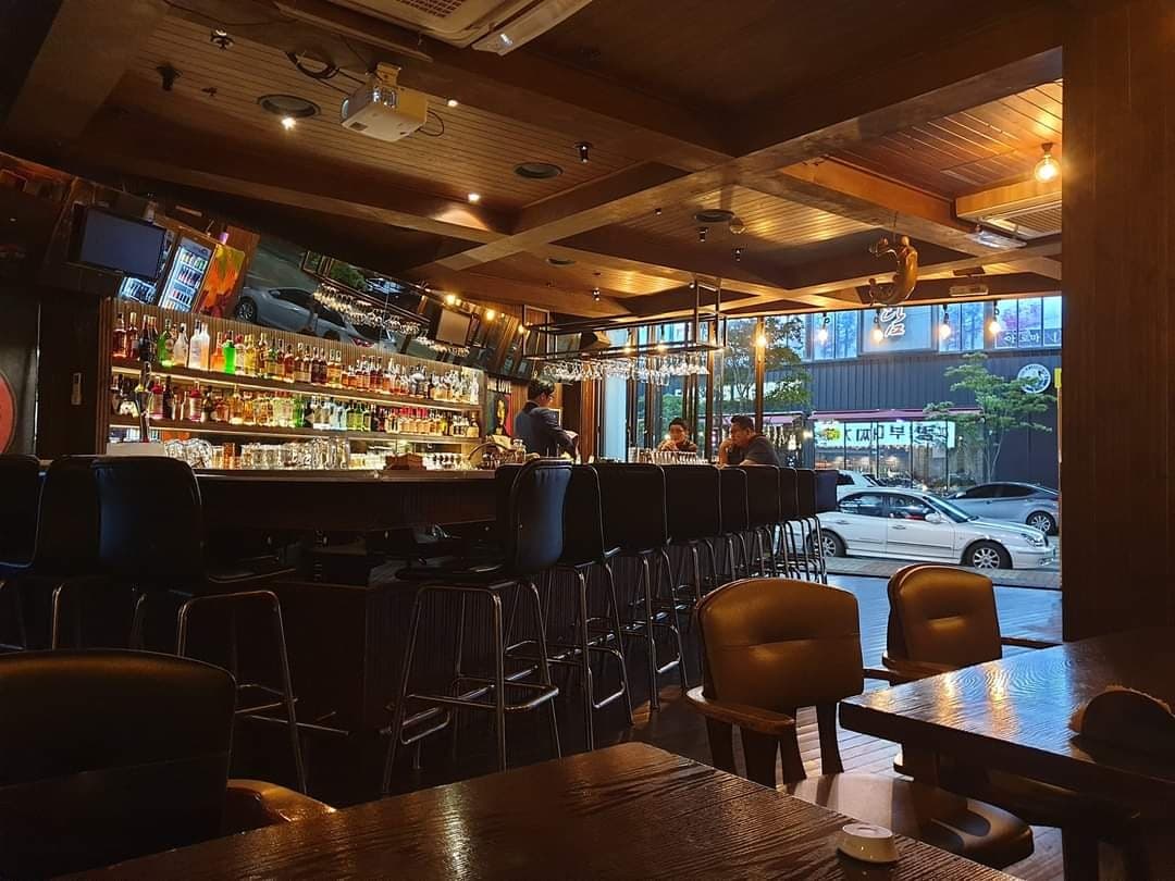 은은한 황색 조명 아래 길게 뻗은 나무 카운터와 일렬로 배치된 높은 의자들, 벽면을 가득 채운 양주병 진열장