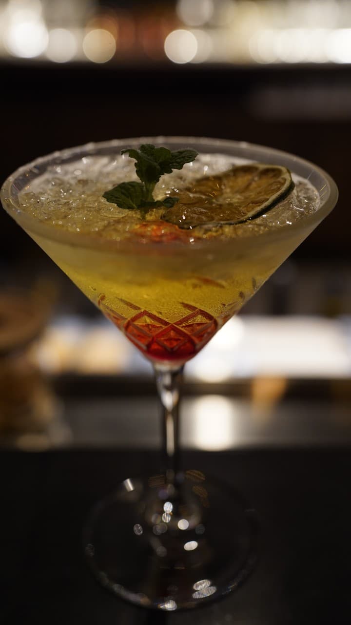 어두운 조명 아래 바 테이블 위 칵테일 한 잔. 잘게 부순 얼음과 말린 라임, 민트 장식, 하단의 붉은 시럽과 배경의 흐릿한 조명.