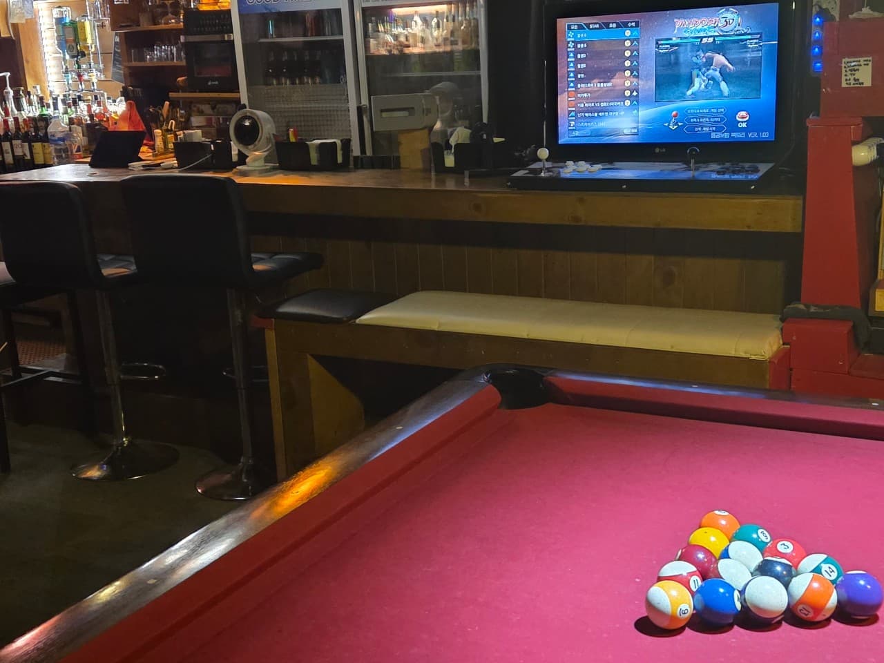 붉은색 당구대 뒤편으로 1인용 의자가 나란히 놓인 긴 목재 카운터와 게임 화면이 켜진 모니터 전경