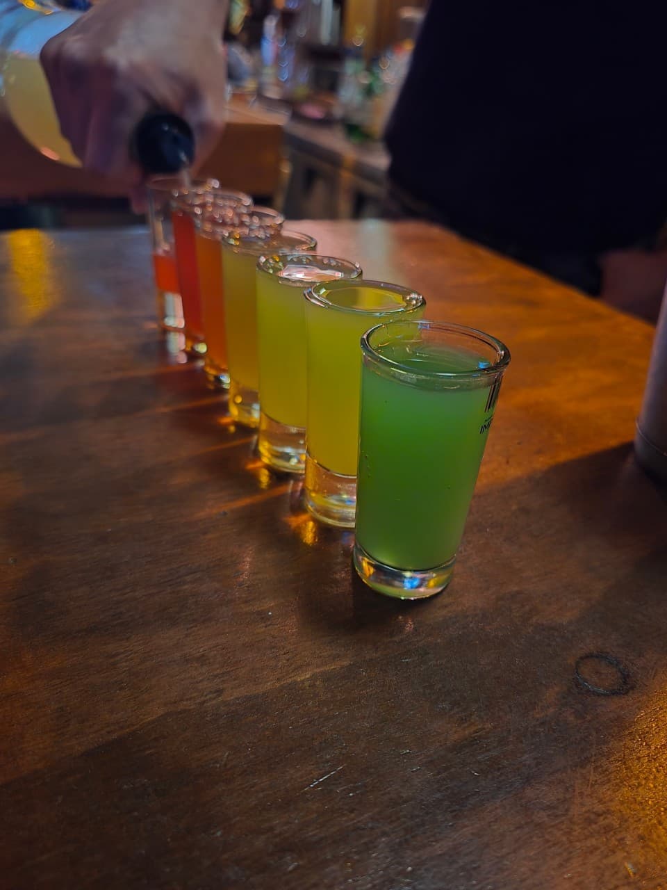 어두운 조명의 나무 테이블 위 일렬로 놓인 다양한 색상의 샷 잔들과 술을 따르는 손