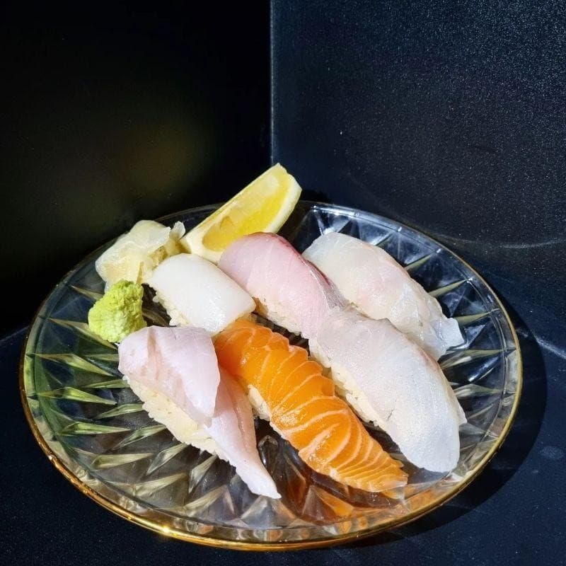 검은 배경 아래 조명을 받은 금색 테두리 유리 접시 위 연어와 흰살생선 초밥 6피스 및 레몬과 고추냉이 곁들임