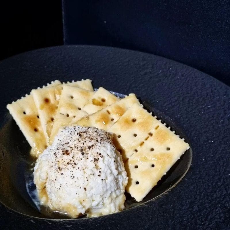 어두운 배경 속 검은색 질감의 그릇에 담긴 꿀이 뿌려진 사각 크래커와 후추를 얹은 크림치즈