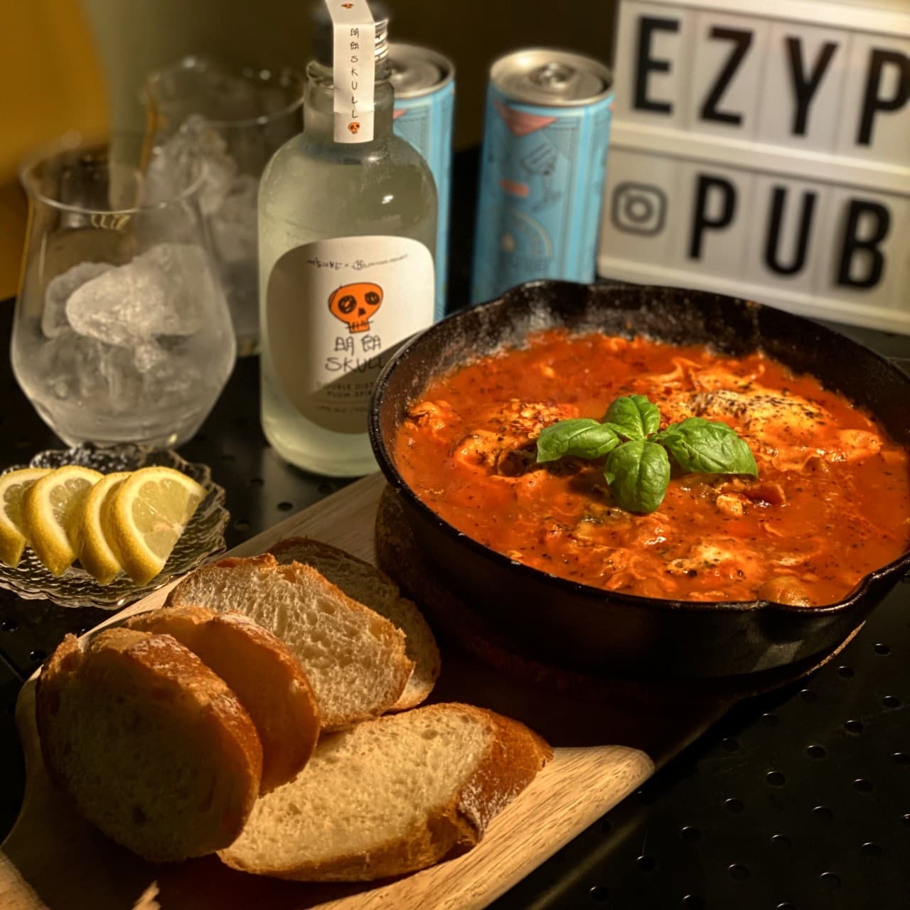 어두운 조명 아래 무쇠 팬 속 토마토 요리와 바질, 나무 도마 위 빵과 술병이 놓인 1인용 테이블