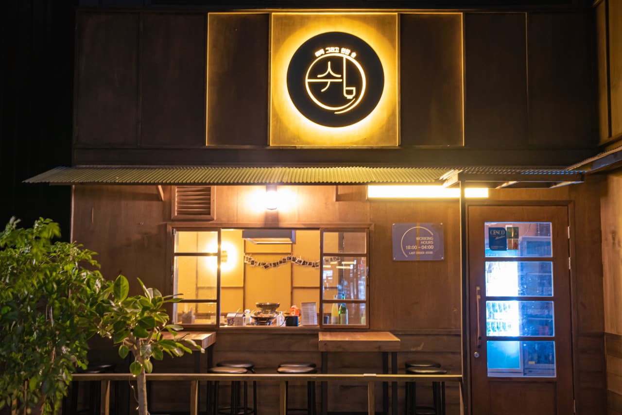 노란 원형 조명 아래 목재 외벽과 야외 카운터석이 배치된 공간. 창가 바 테이블을 따라 놓인 1인용 검은색 원형 의자들.