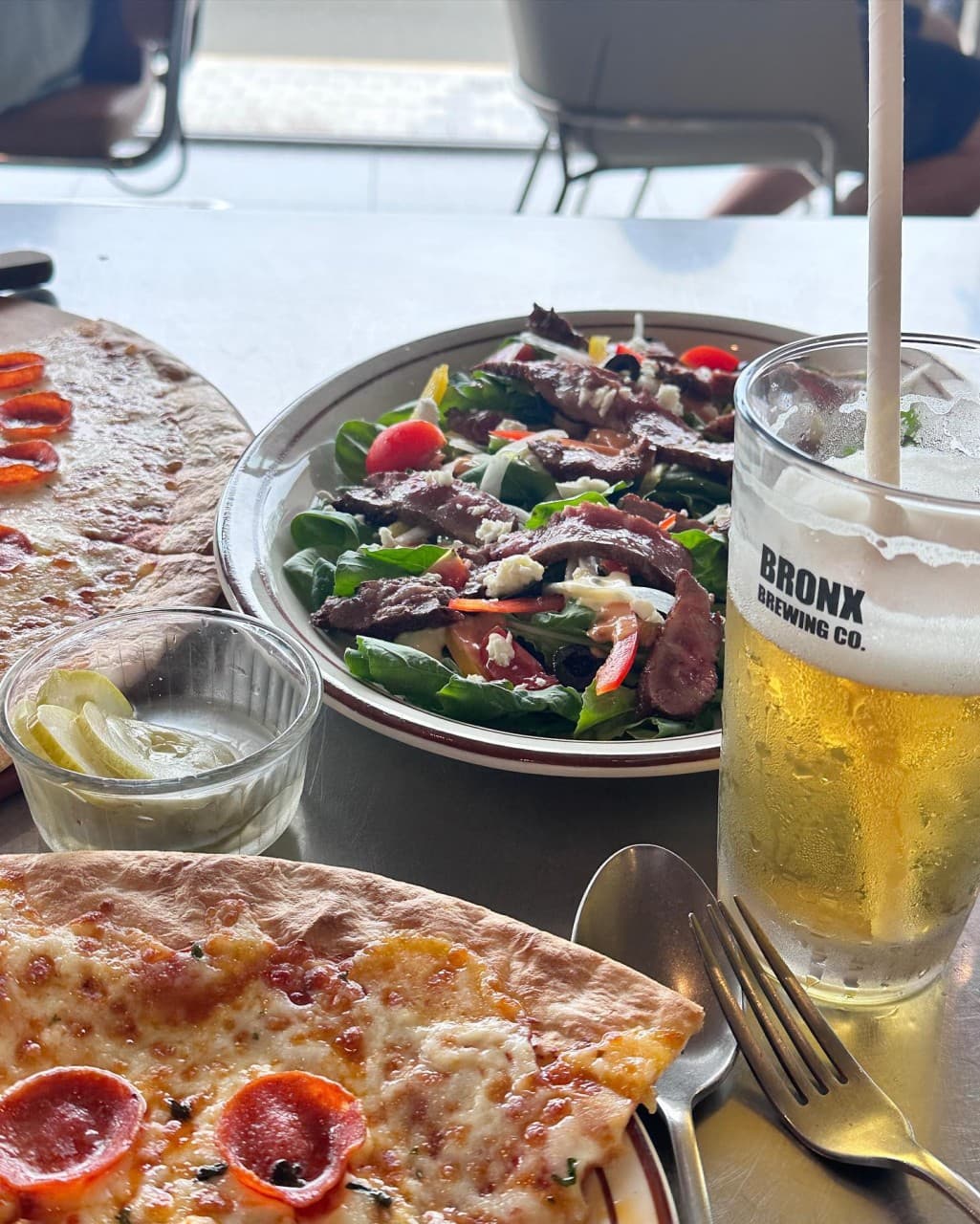 밝은 채광의 금속 테이블 위 페퍼로니 피자와 소고기 샐러드, 거품이 올라온 맥주 한 잔과 1인용 식기류